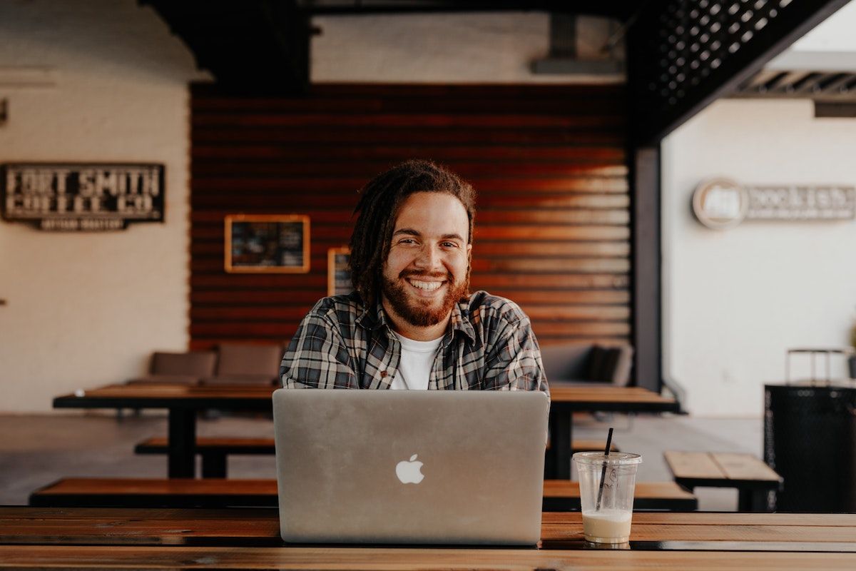 smiling student sitting at a table behind with a MacBook Pro and an iced coffee