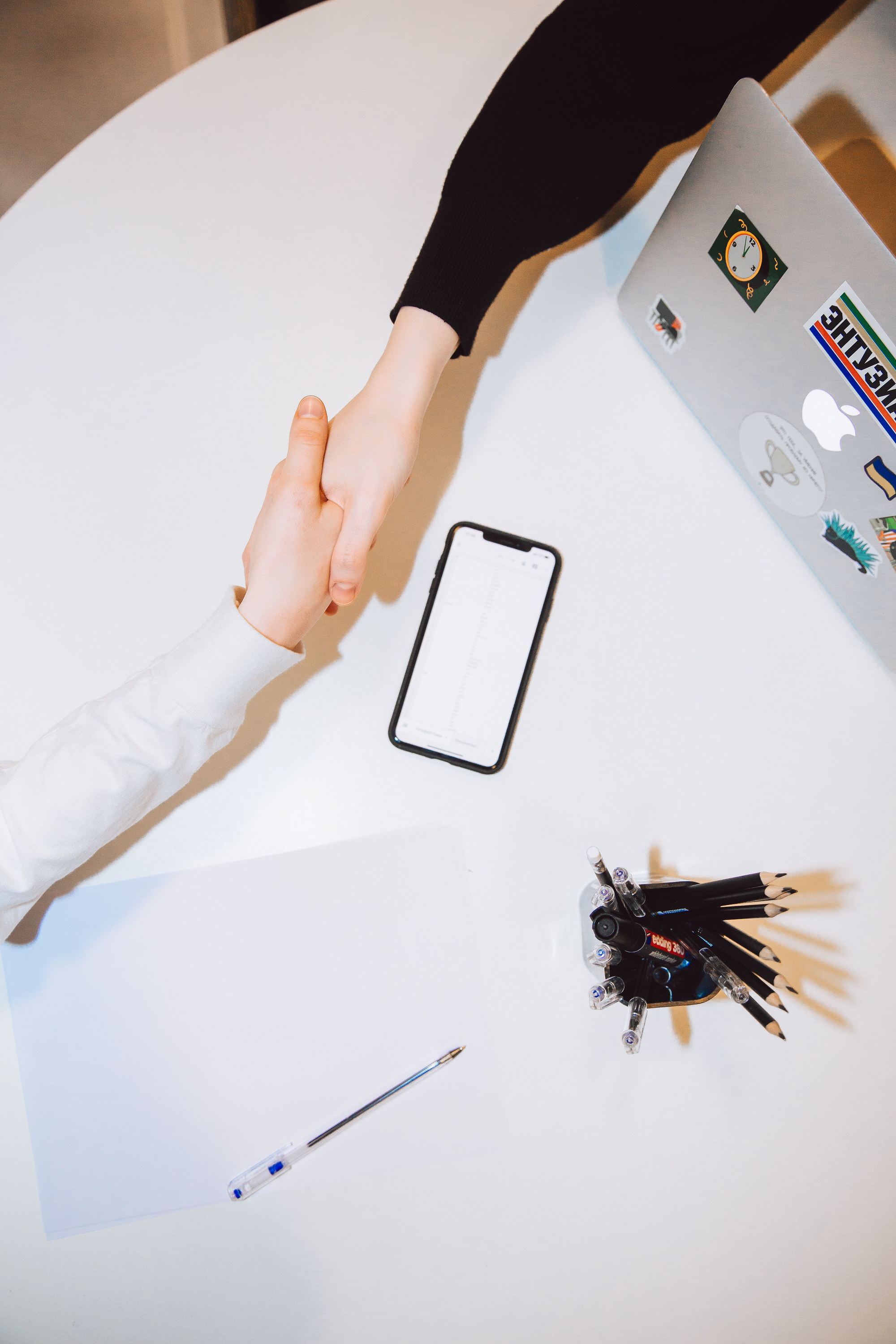 Two hands shaking from above with view of table, smartphone, a pencil jar, and a laptop